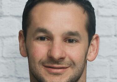 Portrait of a man with short dark hair in front of a gray brick wall.