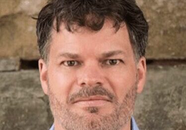 Man with curly dark hair and beard against a stone background.