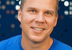 Portrait of a smiling man in a blue shirt with a dotted background.