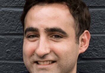 Smiling young man with dark hair in front of a dark wall.