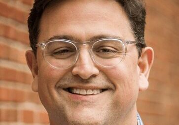 A smiling man wearing glasses and a checkered shirt.