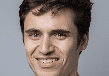 A young man with short brown hair smiling against a plain background.