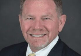 A middle-aged man in a suit smiling against a dark background.
