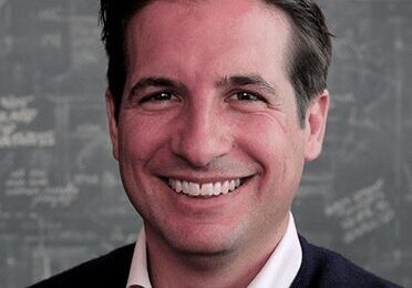 Smiling man in business casual attire against a chalkboard background.
