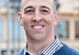 Smiling bald man wearing a blue sweater and checkered shirt outdoors.