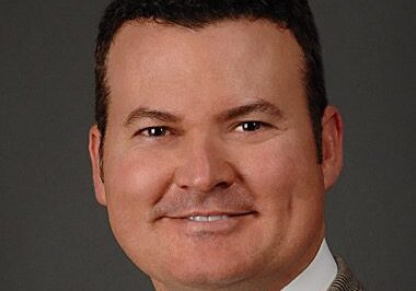 Professional headshot of a man in a beige suit and white shirt.