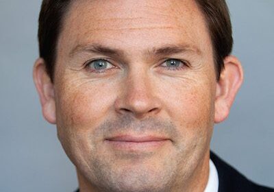 Professional headshot of a confident man in a suit against a gray background.