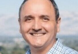 Smiling man in a checkered shirt outdoors with a blurred background.