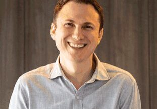 Smiling man in a light blue shirt against a wooden background.