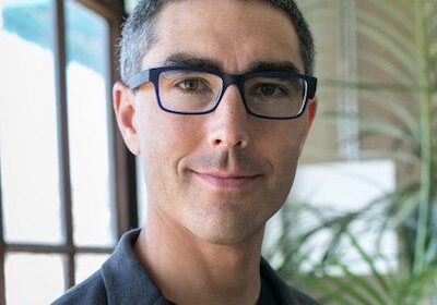 Man wearing glasses and a blazer smiling indoors near a window.