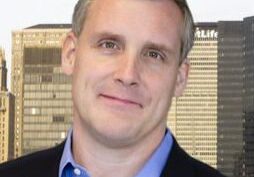 Portrait of a man in a blue shirt and black jacket with a cityscape background.
