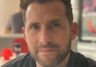 Close-up of a man with a beard and styled hair in a casual setting.
