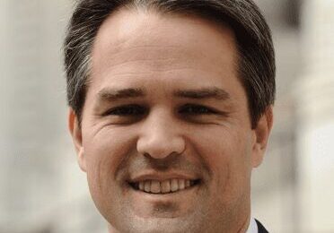 Smiling man in a suit, outdoors with blurred background.