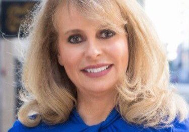 A woman with blonde hair smiling in a blue top.