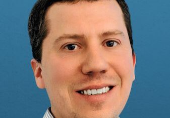 Professional headshot of a smiling man with short dark hair against a blue background.