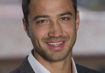 Smiling man in a suit jacket with a blurred background.