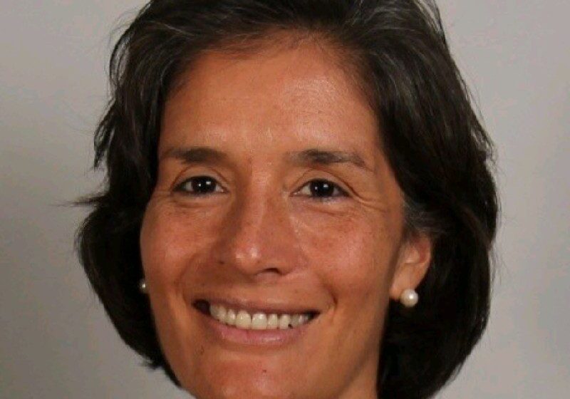 Smiling woman with short dark hair and pearl earrings against a plain background.