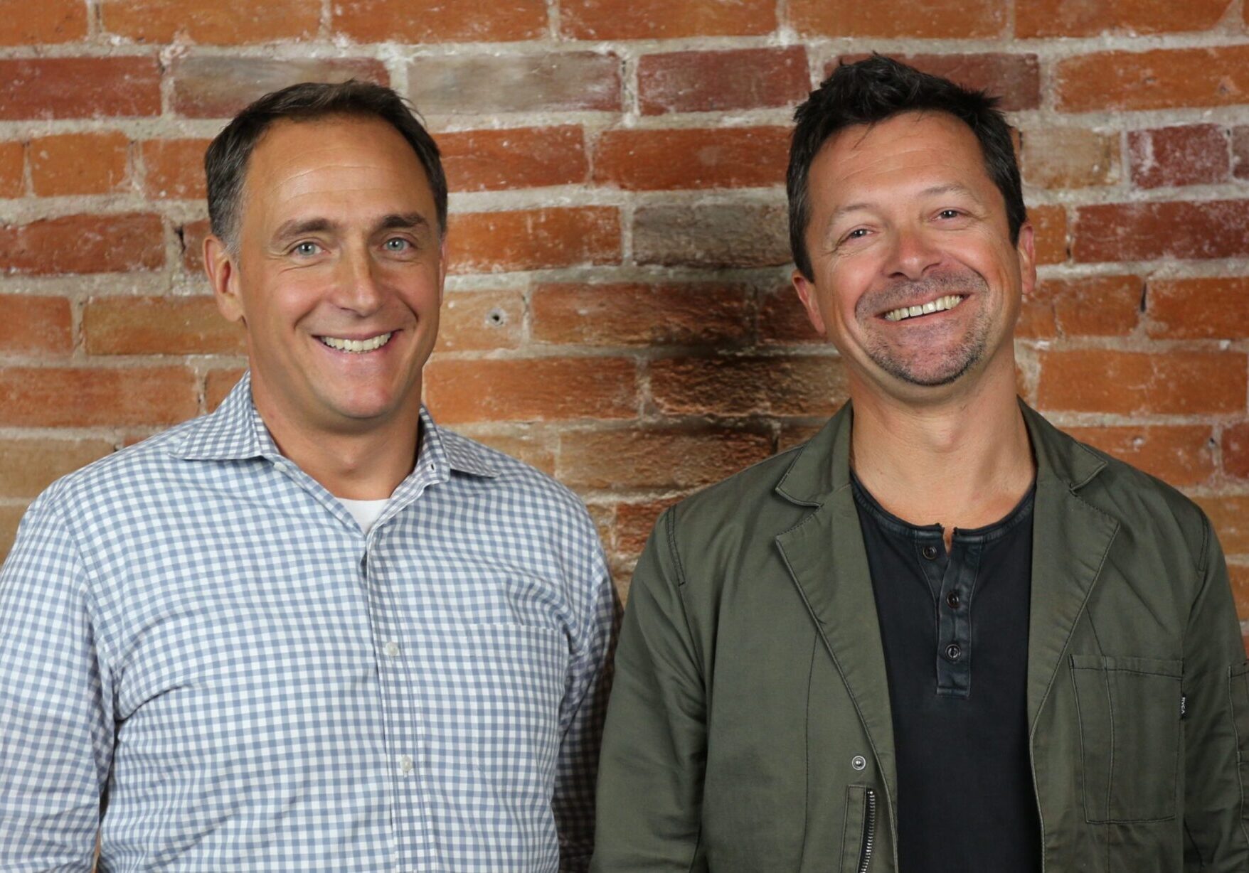 Two men smiling in front of a brick wall background.