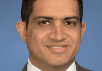 Professional headshot of a man in a suit and tie against a blue background.