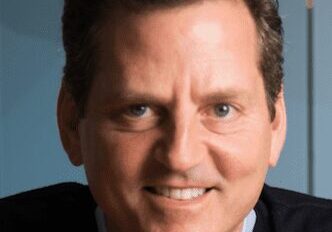 Portrait of a smiling man in a dark jacket with a blue background.