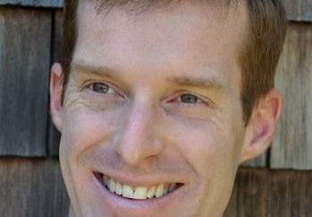 Smiling man with light brown hair in front of a wooden background.