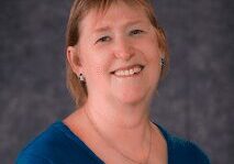 Smiling woman in a blue top against a gray backdrop.