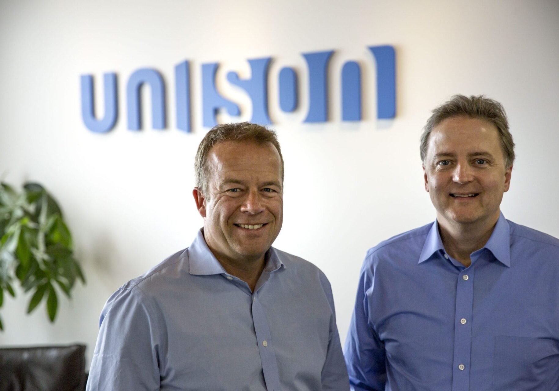 Two men smiling in front of a blurred company logo.