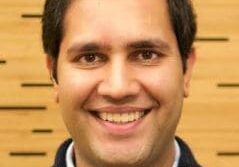 Smiling man with dark hair wearing a navy blue sweater and white collared shirt.