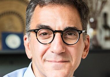 A man with glasses smiling warmly in an indoor setting.